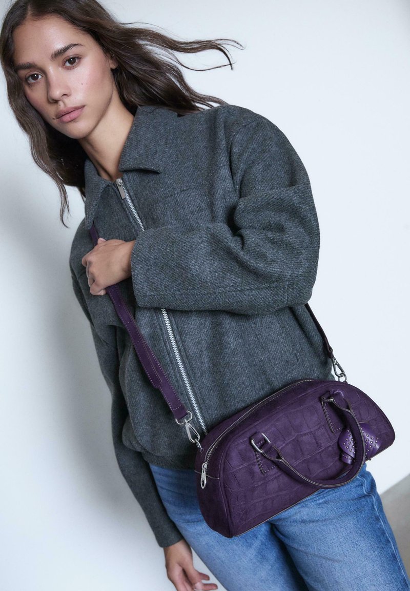 Lila Lederhandtasche mit strukturiertem Krokodilmuster, doppelten Griffen und einem abnehmbaren Schultergurt, kombiniert mit einer grauen Jacke mit Reißverschluss.