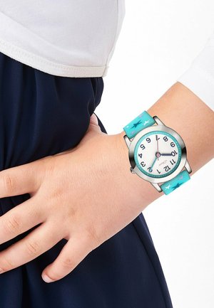 Child's wrist wearing a silver watch with a white dial, black numbers, and blue strap decorated with stars, hand resting on dark blue pants.