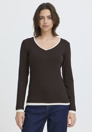 Mladá žena s hnedými vlasmi po ramená, oblečená v čiernom tričku s dlhými rukávmi a výstrihom do V s bielym lemom a modrými džínsami, stojaca na neutrálnej pozadí.