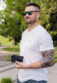 Black zippered wallet held in a hand, with a plain white V-neck shirt and vibrant, reflective sunglasses. Green trees in the background.