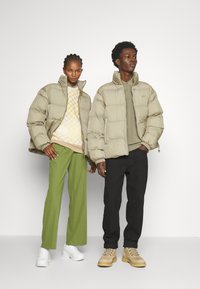 Chaquetas acolchadas beige con cuellos altos. Mujer con un suéter estampado y pantalones verdes, hombre con una camisa verde oliva y pantalones negros. Zapatos blancos y color beige.