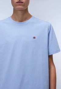 Light blue cotton t-shirt with short sleeves and a round neckline, featuring a small embroidered design in the upper left corner.