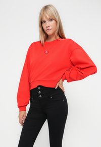 Sweat-shirt rouge court à manches longues, avec une épingle en forme de cœur sur la poitrine ; associé à un jean noir taille haute avec des détails de boutons.