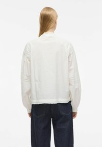 Witte katoenen blouse met lange opgezette mouwen, kantdetails op de schouders en mouwen, en een golvende zoom. Draag met donkerblauwe jeans.