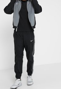 Osoba oblečená v černých Nike teplákových kalhotách s bílými pruhy, černém tričku s dlouhým rukávem, šedé a černé bundě a bílých Nike teniskách.