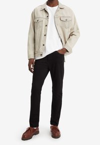Chaqueta de mezclilla gris claro con dos bolsillos frontales, camiseta blanca, jeans negros y zapatos marrones con cordones. Conjunto informal con colores neutros.