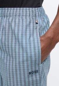 Main dans la poche d'un pantalon à taille élastique à carreaux bleu clair avec une petite étiquette de marque et le logo "BOSS" près de la poche.