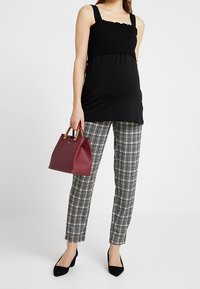 Svart smockad topp med breda band, i kombination med rutiga byxor. Håller en bordeauxfärgad handväska med bruna handtag och har på sig svarta klackar.