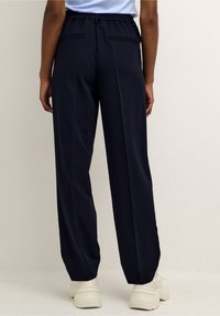 Pantalon bleu marine à coupe décontractée avec une taille élastique, deux poches arrière et une texture lisse, associé à des baskets de couleur claire.