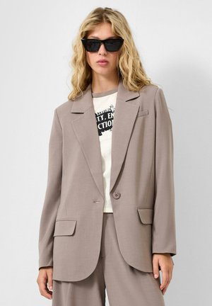 Femme blonde portant une veste de costume beige oversize, un pantalon assorti, un t-shirt blanc à motif graphique et des lunettes de soleil rectangulaires noires, debout devant un fond blanc.