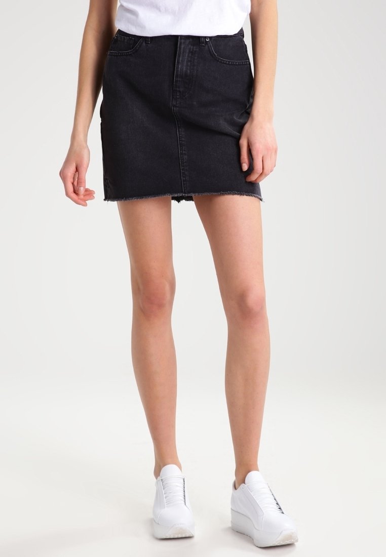 Personne portant un t-shirt blanc, une mini-jupe en jean noire à ourlet effiloché et des baskets blanches, debout devant un fond uni clair.