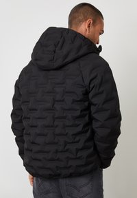 Zwarte pufferjack met een capuchon, met een textuur en een gewatteerd patroon. De achterkant heeft een getailleerd silhouet en elastische manchetten.