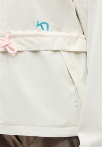 Κοντινή λήψη κρεμ μπουφάν με μπλε λογότυπο, ροζ κορδόνι στην αριστερή πλευρά και κεκλιμένη τσέπη με ανοιχτό μπλε τράβηγμα στη δεξιά πλευρά.