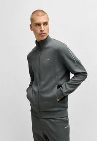Giacca grigia con zip realizzata in tessuto strutturato, colletto alto, due tasche laterali e logo ricamato "BOSS" in oro sul petto e sulla gamba.