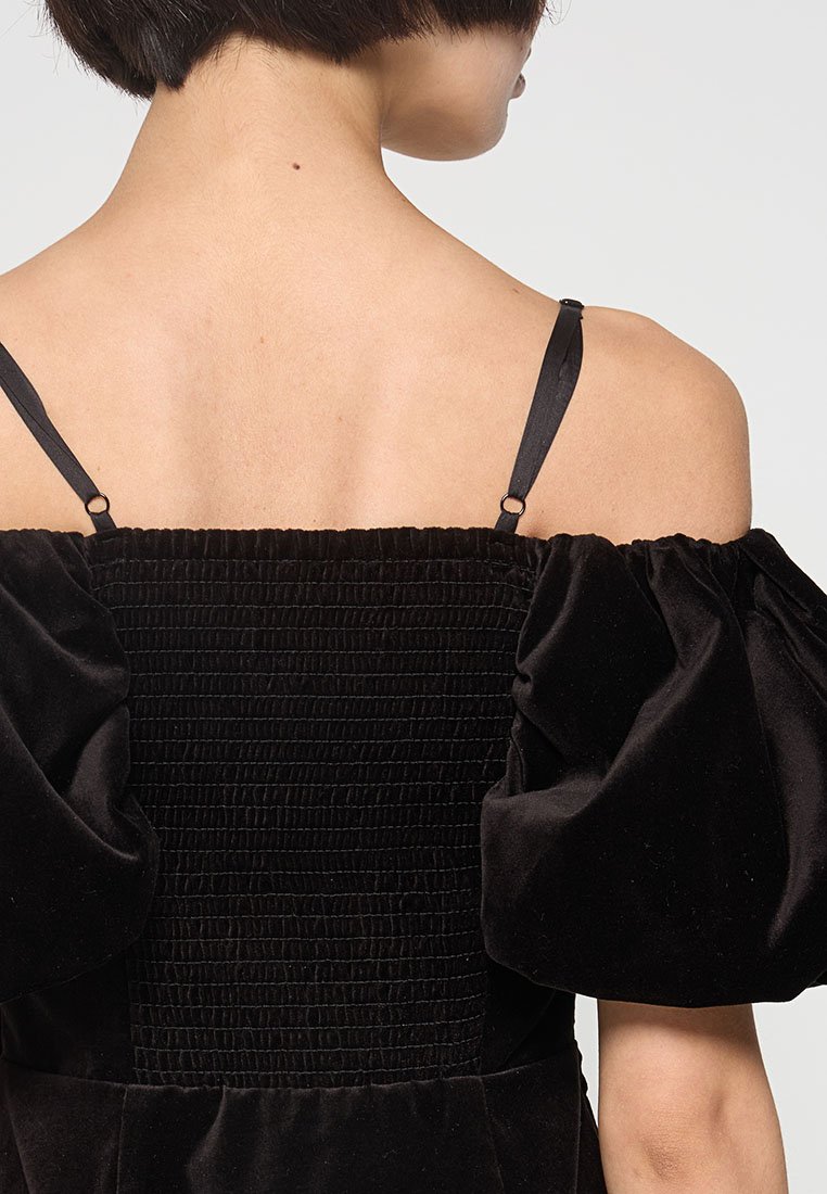 Robe en velours noir avec des manches bouffantes, un dos smocké et de fines bretelles en satin. La texture est douce, avec un design structuré dans l'ensemble.