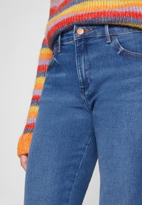 Mørkeblå jeans med en tettsittende design og en kobberknapp i linningen. Tøyet har en jevn tekstur med minimal falming.