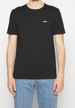 Homme portant un T-shirt noir à manches courtes avec un petit logo blanc "BOSS" sur la poitrine, associé à un jean bleu clair, debout devant un fond blanc.