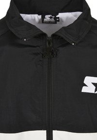 Giacca a vento nera con chiusura a zip, foderata in mesh e logo bianco prominente. Presenta una texture liscia e un design con colletto alto.