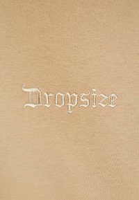 Light brown fabric featuring embroidered text "Dropsize" in white Gothic-style font. Smooth, even texture with no visible patterns.