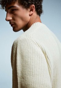 Strickpullover in Cremefarbe mit geripptem Muster, ausgestattet mit einem runden Ausschnitt und dezenten Schulterdetails. Hochformat.