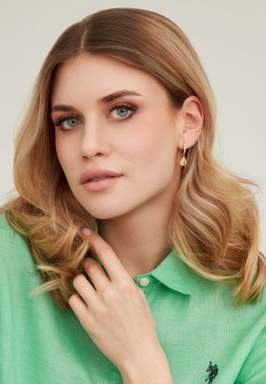 Giovane donna con capelli biondi ondulati che indossa una camicia verde, orecchini d'oro a forma di goccia e si tocca delicatamente il collo.