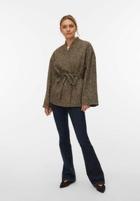 Brūna zivju kaulu raksta wrap jakas ar jostu, garām piedurknēm un vaļēju piegriezumu, kombinēta ar tumšām zvīņainām džinsu biksēm un norādītām apavu pirkstu daļām.