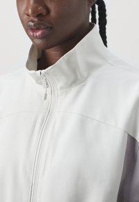 White zip-up jacket with high collar, featuring a smooth texture and a contrasting gray panel on the right side. Subtle stitched detailing.