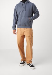 Felpa grigia con tasca a canguro e testo "NEW BALANCE 1906". Pantaloni cargo beige con tasche laterali. Sneakers in bianco e nero.
