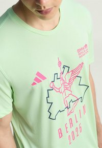 Camiseta atlética verde claro con un diseño gráfico de una figura alada y el contorno del mapa de Berlín, acentuada con elementos en rosa y azul oscuro.