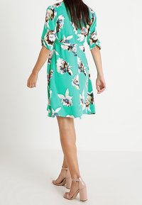 Abito floreale verde con maniche a sbuffo corte e cintura, caratterizzato da un orlo fluido. Indossato con sandali con tacco a blocco rosa chiaro.