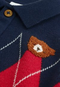 Gros plan sur un pull bleu marine avec un motif argyle rouge et blanc et un détail de visage d'ours en laine tricotée près du col à bouton en bois.