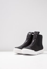 Černé sneakers do kotníků s texturovanými síťovanými panely, embosovaným brandingem a silnou bílou gumovou podrážkou. Obsahují design pro snadné obutí a polstrovaný límec.