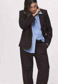 Brązowa, dopasowana marynarka na niebiesko-białej koszuli w paski, z ostrym kołnierzykiem, zapinana na guziki, w połączeniu z luźnymi, czarnymi spodniami.