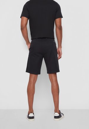 Schwarze Shorts mit maßgeschneiderter Passform, gefertigt aus leichtem Stoff. Verfügt über zwei Gesäßtaschen und eine glatte Textur. Kombiniert mit schwarzen Sneakers.