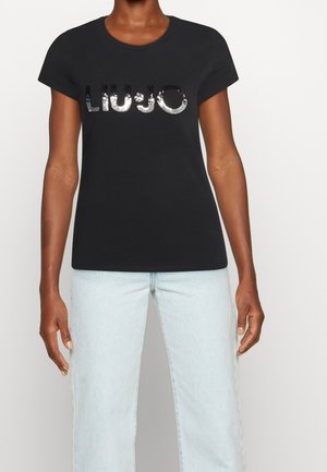 Person wearing black short-sleeve t-shirt with shiny sequined "LIU JO" logo and light blue jeans, standing against a plain white background.