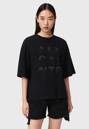 Femme aux cheveux raides et foncés portant un T-shirt noir oversize avec le texte clouté "ALL SAINTS" et un short en denim noir effiloché.
