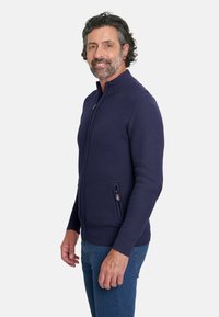 Suéter de punto con cremallera en azul marino oscuro. Presenta un cuello alto, un patrón texturizado y bolsillos laterales. Lleva puesto unos vaqueros azules.