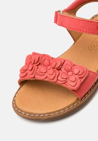 Korallfärgade läder sandaler med blommor på remmen, tanfärgad innersula och brun yttersula; har justerbar ankelrem.