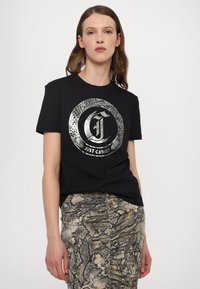 T-shirt en coton noir avec un logo argenté brillant et des motifs de serpent. Encolure ronde simple et manches courtes, associé à une jupe à motifs.