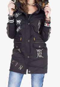 Parka nera con cappuccio e bordo in pelliccia, dotata di tasche, toppe decorative con testo, dettagli borchiati e chiusura con bottone.