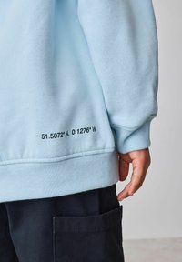 helehall sinine fliistekstuuriga pusa, millel on musta värvi trükitud koordinaadid äärel. Kandmisel tumedate pükste kohal, millel on küljetasku.