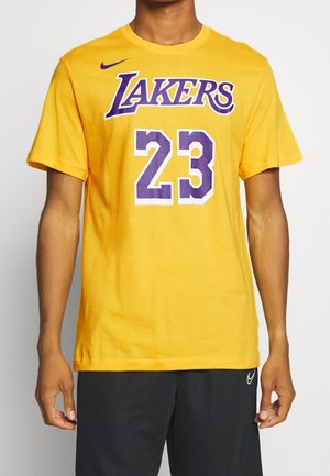 Osoba nosząca żółtą koszulkę Los Angeles Lakers z numerem 23 w kolorze fioletowo-białym, połączoną z czarnymi szortami Nike.