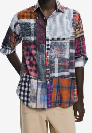 Koszula patchwork z długim rękawem z różnymi wzorami w kratę i paski w kolorach pomarańczowym, czarnym, białym i niebieskim, noszona z beżowymi spodniami.