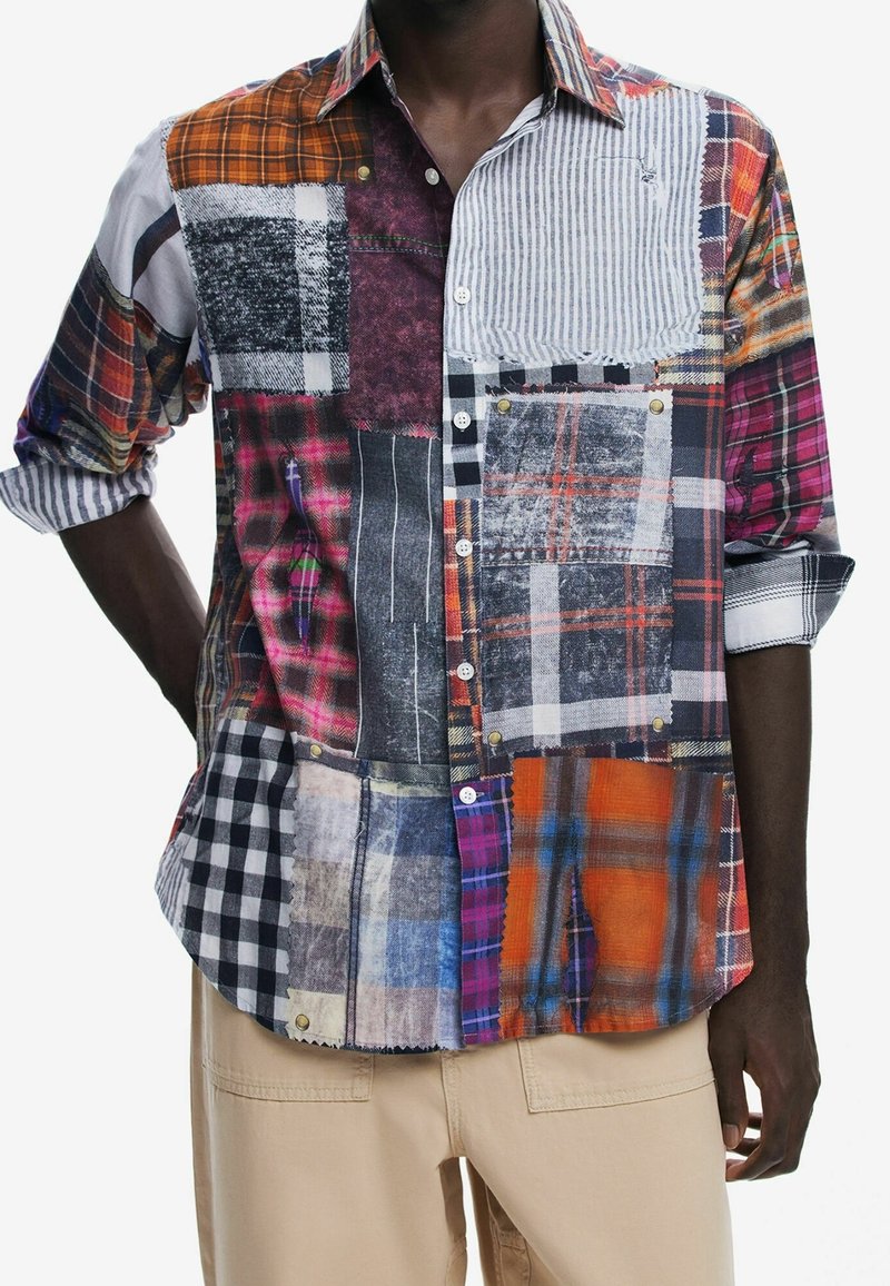 Chemise patchwork à manches longues avec différents motifs à carreaux et rayures en orange, noir, blanc et bleu, portée avec un pantalon beige.