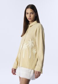 Camisa amarilla texturizada con cuello, adornada con bordado de palmeras blancas en el frente. El modelo lleva pantalones cortos blancos y posa de lado.