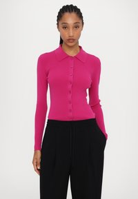 Fuchsia geribde cardigan met lange mouwen, knoopsluiting aan de voorkant en opstaand kraag. Gecombineerd met zwarte broeken met een koord in de taille.