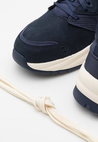 Mörkblå mockaskor med vit sula bredvid en knuten ljusbeige skosnör på en vit bakgrund.