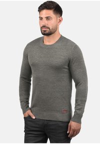 Grijze gebreide trui met lange mouwen, ronde hals en ribgebreide manchetten. Heeft een klein logo-patch nabij de zoom. Glad textuur.