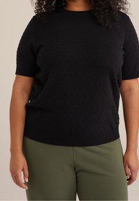 Kvinna iförd en svart texturerad kortärmad tröja och olivgröna byxor, sedd från halsen till höfterna mot en enfärgad bakgrund.