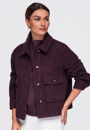 Femme portant une veste en laine violet foncé boutonnée, avec de grandes poches avant et un col plié, regardant sur le côté avec une main sur la hanche.